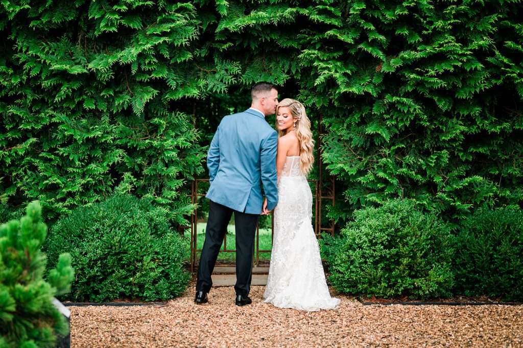 Groom kissing bride's head in front of greenery outside