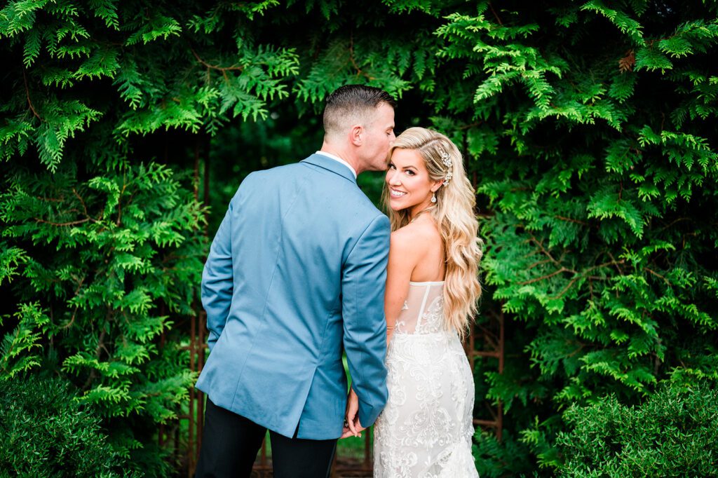 Groom kissing brides head with greenery in background